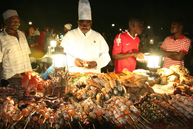 Cooks at a night food stall lit by lanterns, skewers of seafood on the grill