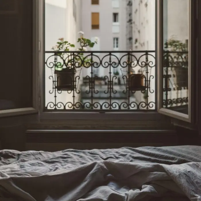 An unmade bed facing tall open shutters and a wrought-iron balcony with potted plants