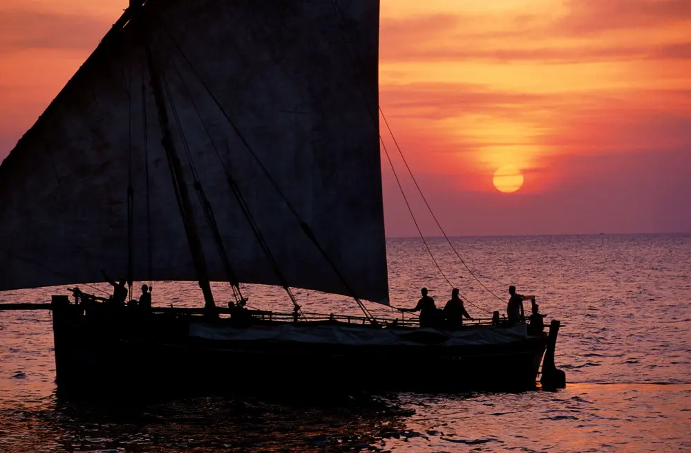 A dhow under full sail silhouetted against a red sun setting into the sea