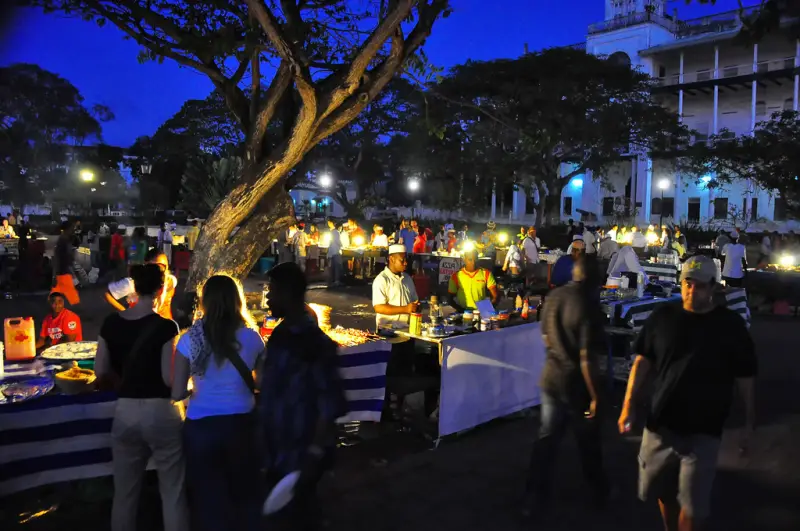 A busy night food market under trees, lit by lamps